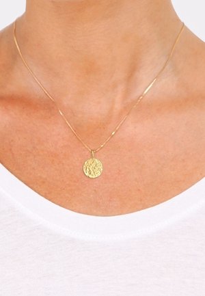 Pendentif rond texturé en or sur une fine chaîne en or, porté autour du cou d'une personne avec un haut blanc.