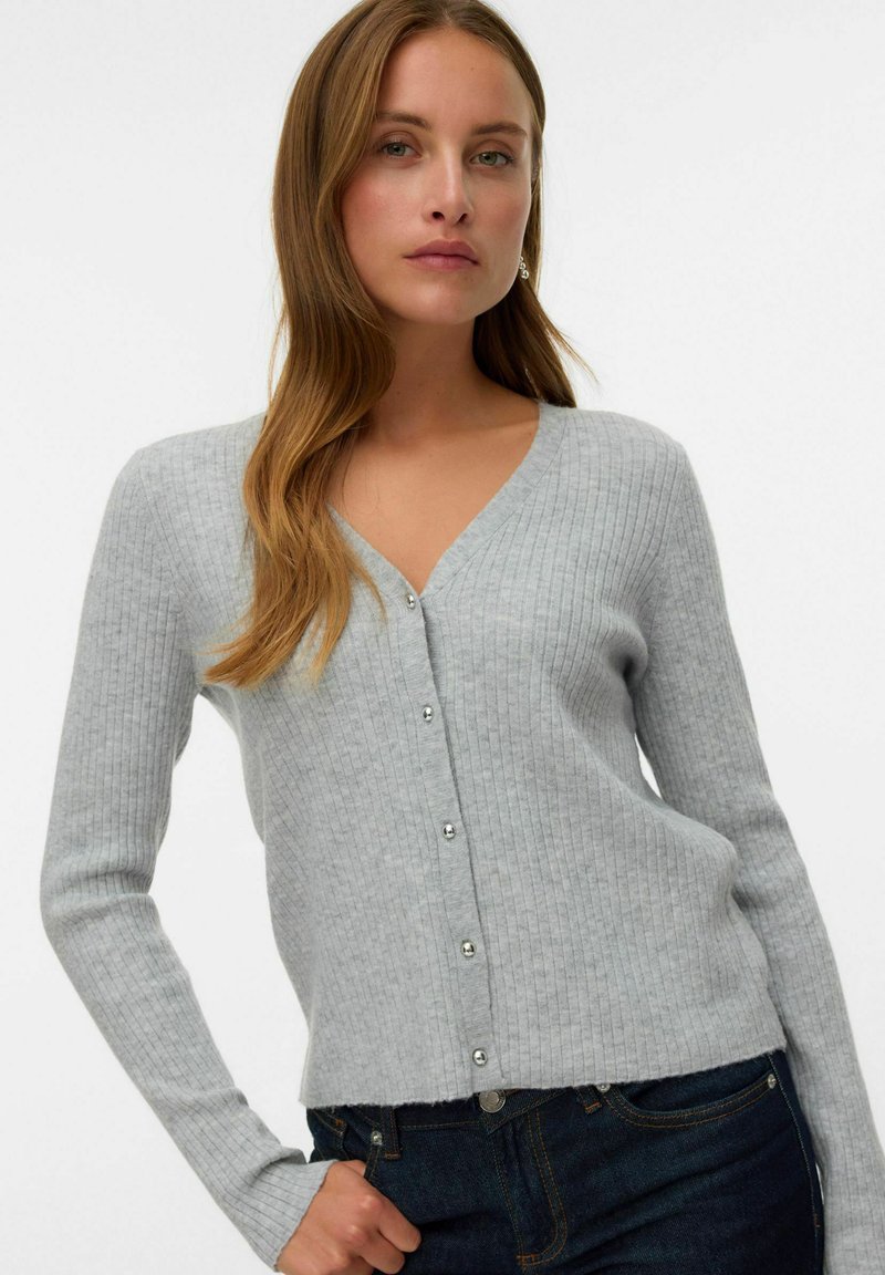 Grijze geribbelde cardigan met een V-hals, lange mouwen en knopen aan de voorkant. Het materiaal lijkt zacht met een lichte structuur. Draagt over donkere jeans.
