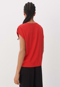 Rotes Kurzarmoberteil mit lockerem Sitz, glatter Textur und rundem Ausschnitt. Verziert mit einem dezenten Cut-out an den Schulternähten.