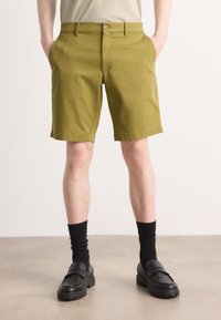 Olivengrønne bomullsshorts med sidelommer, knappelukking og rett passform, paret med svarte sokker og loafers.