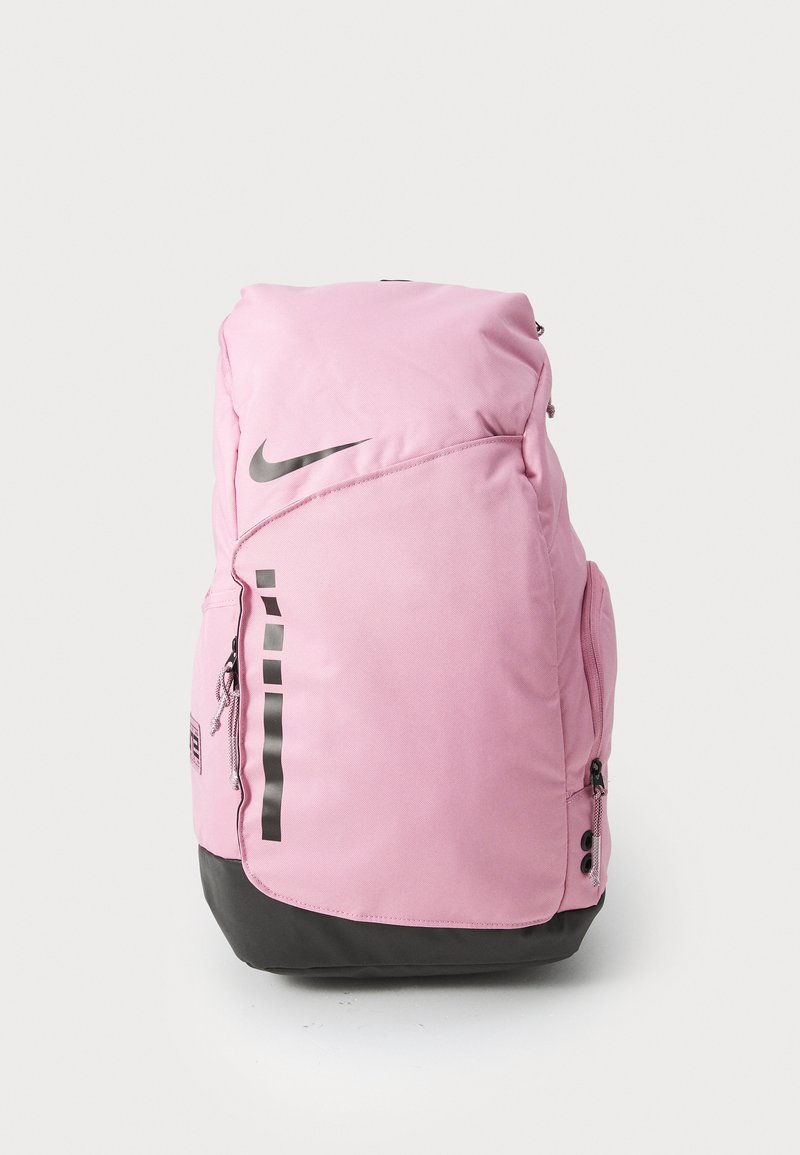 Rozā neilona mugursoma ar melnu apakšu, kurai ir liels Nike logotips, sānu rāvējslēdzēja kabata un vertikālas svītras uz priekšpuses.
