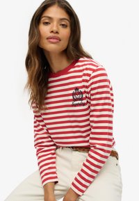 Rood-wit gestreepte langarmshirt met een ronde halslijn, voorzien van een geborduurd logo. Gepaarde met beige broeken en een gevlochten riem.