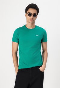 Green short-sleeve t-shirt made of cotton, featuring a round neck and a small white logo on the chest. Model wears black pants and sunglasses.