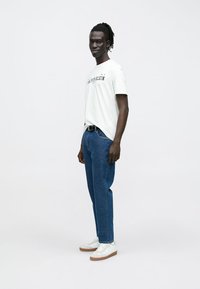 Balta kokvilnas T-krekls ar melnu logotipu, zili džinsa bikses un balti sporta apavi ar brūnām zolēm. Vienkāršs un ikdienišķs siluets.