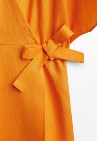 Tissu en lin orange noué en nœud à la taille, créant des fronces subtiles et une texture sur un vêtement sur fond blanc.