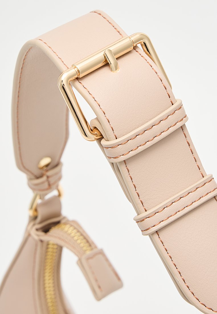 Tracolla in pelle beige con fibbia rettangolare dorata e dettagli cuciti, attaccata a una borsa con una cerniera dorata visibile.
