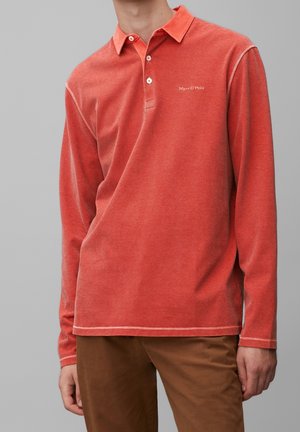 Mężczyzna w długim rękawie, wyblakłej czerwonej koszulce polo z cienkimi, poziomymi paskami oraz brązowych spodniach na jednolitym, szarym tle.