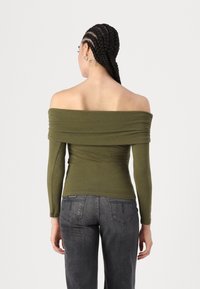 Oliven grønn off-the-shoulder topp med lange ermer, rynkede detaljer over bysten, kombinert med mørkegrå jeans. Glatt, tettsittende tekstur.