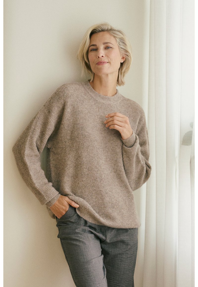 Femme blonde en pull en tricot beige et pantalon gris, debout, détendue, près d'un rideau blanc, avec une main dans la poche et un léger sourire.