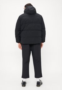 Black puffer jacket with a hood, featuring horizontal quilted design. Matte finish and loose fit. Worn with black pants and shoes.