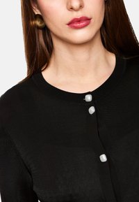 Schwarze Strickjacke mit geripptem Muster, ausgestattet mit drei runden, glänzenden weißen Knöpfen. Nahaufnahme, die den Kragen und die Knopfdetails betont.