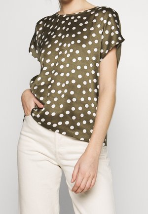 Femme portant un haut à manches courtes vert olive à pois blancs et un pantalon taille haute écru, une main dans la poche.