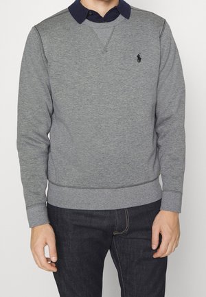 Mann trägt ein graues langärmeliges Sweatshirt über einem dunkelblauen Hemd mit Kragen und dunklen Jeans, steht vor einem weißen Hintergrund.