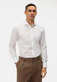 Witte blouse met lange mouwen, een puntkraag, knoopsluiting en een gladde textuur, gecombineerd met een bruine broek met een geplooide voorkant.