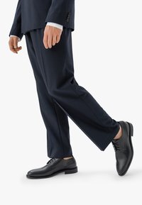 Scarpe eleganti in pelle nera con texture liscia e design con lacci, abbinate a pantaloni su misura a righe sottili scuri.