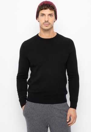 Pullover nero a girocollo realizzato in tessuto morbido, con maniche lunghe e vestibilità aderente, abbinato a pantaloni da casa grigi. Cappellino sulla testa del modello.