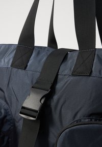 Sac en nylon bleu marine avec des poignées noires, une boucle en plastique solide et une surface texturée avec un motif à carreaux subtil.