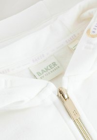 Valkoinen vetoketjullinen huppari, jossa metallinen kultainen vetoketju. Pehmeässä kankaassa ja kuvioidussa vuorissa on "BAKER"-etiketti. Koristeena hienovarainen brändäys.