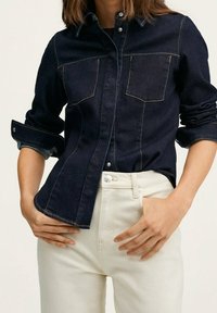 Femme portant une veste en denim bleu foncé aux manches retroussées et un pantalon taille haute blanc cassé, les mains posées sur les hanches.