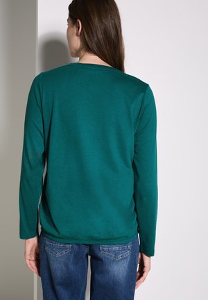 TÜRKIS langärmliges Hemd mit rundem Ausschnitt und elastischem Saum, kombiniert mit blauen Jeans. Der Stoff wirkt glatt mit leichter Textur.