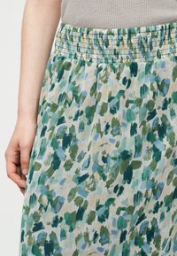 Κοντινό πλάνο ενός ατόμου που φοράει φούστα με ελαστική μέση και αφηρημένο λουλουδόμορφο μοτίβο σε πράσινο, μπλε και μπεζ χρώμα, μαζί με ένα μπεζ ριμπ τοπ.