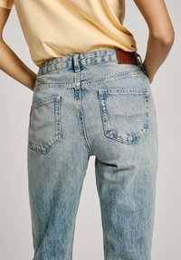 Gai zilo augsto jostu džinsus ar nodilušu tekstūru, kuriem ir divas aizmugures kabatas un brūns ādas plāksteris uz jostas.