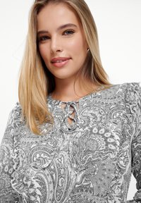Siva bluzica s uzorkom paisley, s okruglim izrezom, detaljem za vezanje i volanima na rukavima. Mekana tkanina naglašava složeni uzorak.