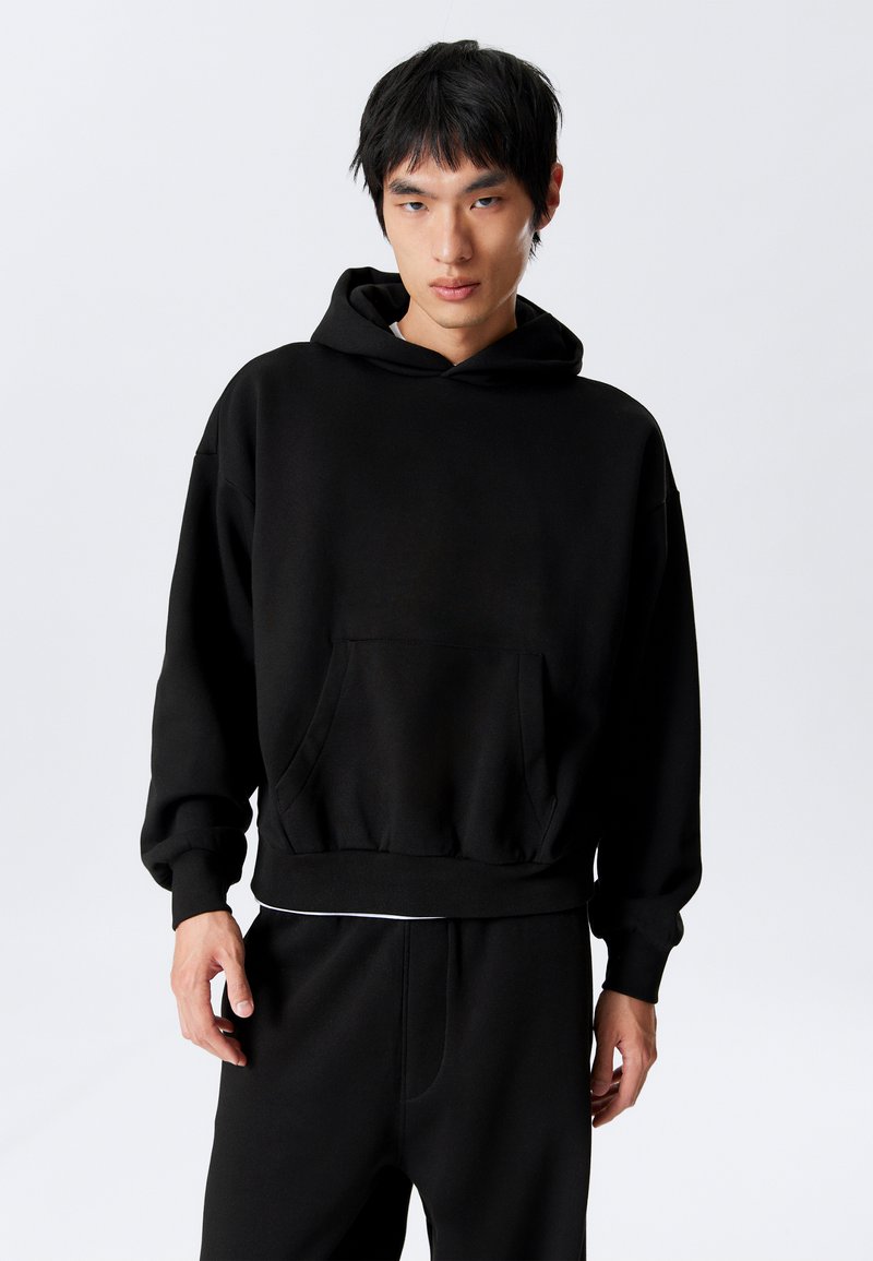 Junger Mann in einem schwarzen Hoodie und passenden schwarzen Hosen, der vor einem schlichten weißen Hintergrund steht.