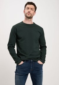 Ciemnozielona koszulka z długim rękawem i okrągłym dekoltem, w zestawieniu z niebieskimi dżinsami. Materiał wydaje się miękki, o prostym, klasycznym wzorze.