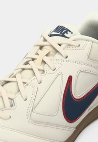 Chaussure de sport blanche avec un dessus en cuir texturé, un swoosh bleu marine et des accents rouges. Les lacets de couleur beige clair complètent le design.