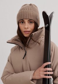 Beżowa kurtka puchowa z wysokim kołnierzem i zamkiem błyskawicznym, z miękką podszewką. Brązowa czapka typu beanie z ribbowe warkocze. Czarne narty z białymi detalami.