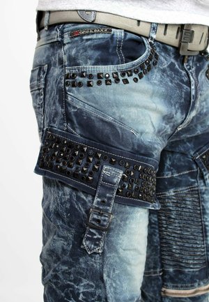 Close-up van vervaagde blauwe spijkerbroek met zwarte met studs versierde bandjes en een grijze riem met een plastic gesp.