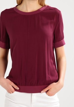 Vrouw draagt een bordeauxroze blouse met korte mouwen en een ronde hals, lichtjes in een witte broek gestopt, handen rustend in de zakken.