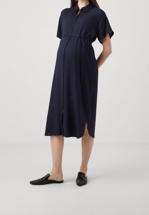 Gravid kvinde iført en marineblå midi-kjole med knapper og bælte i taljen samt sorte slip-on sko, stående foran en ensfarvet baggrund.