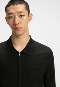 Camicia nera in maglia con zip e colletto, dotata di tasca sul petto e una texture liscia. Inquadratura ravvicinata del capo indossato da un modello.