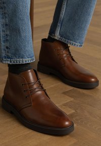 Braune Leder-Ankle Boots mit glatter Oberfläche, runder Zehenpartie, dünnen Schnürsenkeln und schwarzen Gummisohlen. Getragen mit blauen Denim-Jeans.