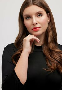 Černý pletený top s vysokým výstřihem. Dlouhé, vlnité hnědé vlasy. Neutrální make-up s výraznými červenými rty. Ruka opřená o bradu.