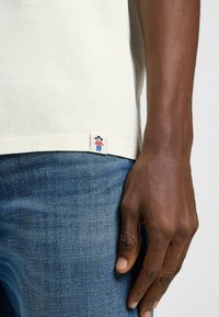 Cremefarvet bomulds-t-shirt med korte ærmer, prydet med et mærke ved kanten, der viser en tegneseriefigur i rød og blå på en hvid baggrund.