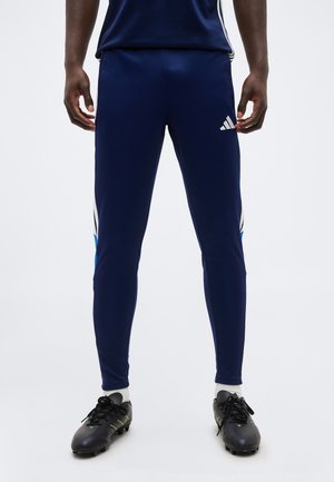 Άνδρας που φοράει παντελόνι φόρμας Adidas σε ναυτικό μπλε και μαύρα παπούτσια ποδοσφαίρου, στέκεται σε απλό λευκό φόντο.