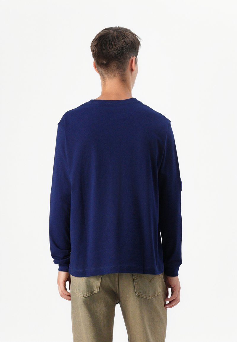 Osoba stoi tyłem do kamery, ubrana w granatową koszulę z długim rękawem i jasne khaki spodnie, na tle jednolitego białego tła.