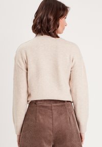 Beige strikket sweater med sænkede skuldre, ribstrikkede manchetter og en subtil tekstur, parret med brune corduroy bukser med høj talje.