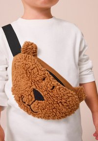 Brun, nallebjörnsformad crossbody-väska i fluffigt tyg, med svarta ansiktsdetaljer och dragkedja; justerbar svart rem.