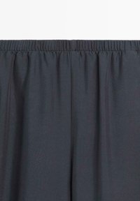 Zwarte broek met elastische tailleband, getoond van de taille naar beneden, met focus op de geplooide stof en naaddetails bovenaan.