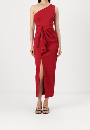 Femme en robe midi rouge à une épaule avec un détail volanté à l'avant et une fente haute sur la cuisse, portant des sandales noires à lanières.