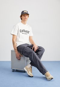 Weißes T-Shirt mit kurzen Ärmeln und schwarzem Text, dunkle Cargo-Hosen und beige Sneakers. Sitzt auf einer silbernen Metallbox mit Rädern.