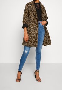 Abrigo de estampado de leopardo con cuello, corte holgado y mangas anchas, combinado con jeans ajustados azules y tacones negros de tiras.