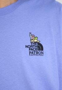Violetinio medvilninio marškinėlių su mažai išsiuvinėtu logotipu, kuriame pavaizduoto šliužo ir kitų elementų, taip pat tekstu "THE NORTH FACE PATRON".