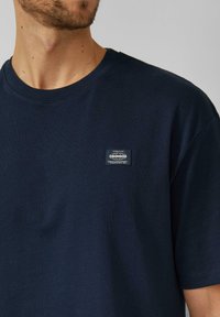Marineblauw katoenen T-shirt met een ronde hals, korte mouwen en een klein stoffen label op de borst met tekst- en logogegevens.