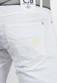 Jean en denim blanc avec des coutures jaunes, présentant un logo en forme de diamant sur la poche arrière et une étiquette de taille grise.