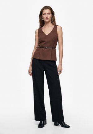 Donna che indossa un top marrone senza maniche con scollo a V e cintura sottile, pantaloni neri a gamba larga e stivali neri con tacco, in piedi davanti a uno sfondo semplice.